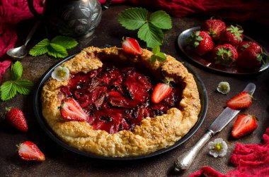 Mayasız hamur çilek pasta için yuvarlak bir şekil ile pasta. Koyu arka plan. Yeşil çilek yaprakları, taze çilek çilek. Kırmızı koşucu. Kırmızı pasta kabuk. Seçici odak. Karanlık fotoğraf.