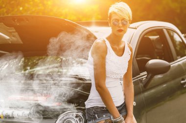 Young sad woman standing near the road by the broken car in the middle of nowhere. Smoke coming out from overheat engine. Waiting car tow service. clipart