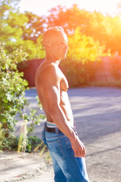 Bellissimo maschio con gli occhi blu e sexy corpo muscoloso indossare jeans di jeans e occhiali da sole in oro con busto nudo. Fitness maschio con abbronzatura sana in posa . — Foto Stock
