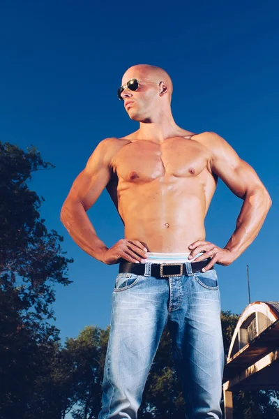 Strong male with blue eyes and sexy muscular body wear denim jeans and gold sunglasses with naked torso. Fitness male with healthy tan posing.
