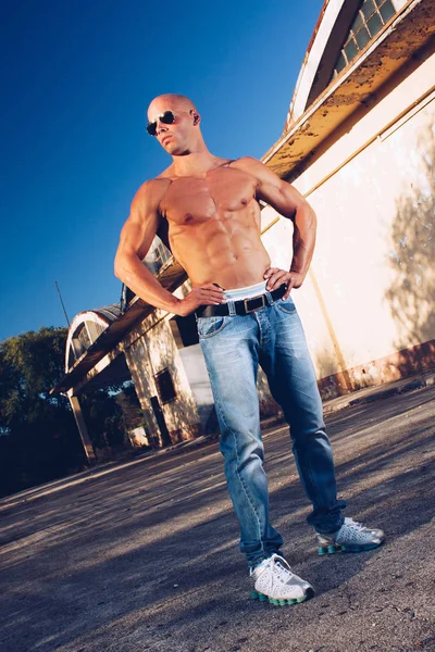 Starker Mann mit blauen Augen und sexy muskulösem Körper trägt Jeans und goldene Sonnenbrille mit nacktem Oberkörper. Fitness-Männchen mit gesunder Bräune posiert. — Stockfoto