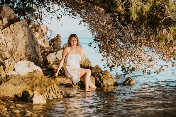 白い濡れたドレスを着た美しい若い妊婦は 夕日のコンセプト愛と妊娠で海水の近くの岩の上でリラックス夏 — ストック写真