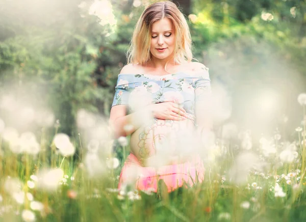 Pregnant woman with mehendi pattern on her belly is sitting on the field with dandelions — Stock Photo, Image