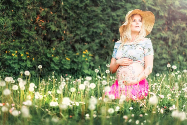 Pregnant woman with mehendi pattern on her belly is sitting on the field with dandelions — Stock Photo, Image