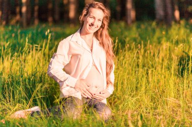 Hamile bir kadın akşam güneşinin ışınlarında ormanın kenarında oturur..