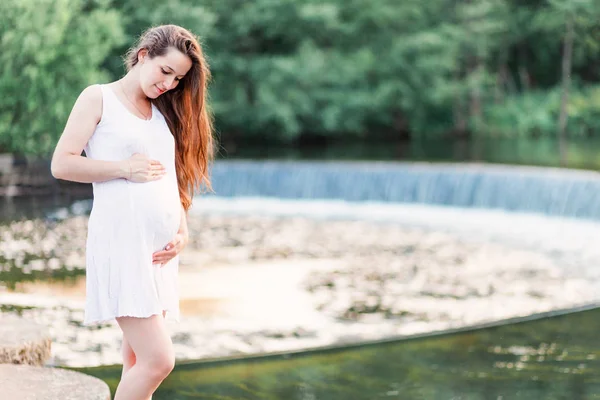 美丽的孕妇站在河边，有一个小瀑布 — 图库照片