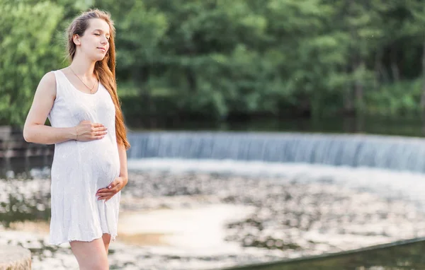 Hermosa mujer embarazada se encuentra cerca del río con una pequeña cascada —  Fotos de Stock