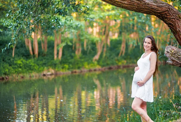 Krásná těhotná žena chodí v parku podél řeky — Stock fotografie