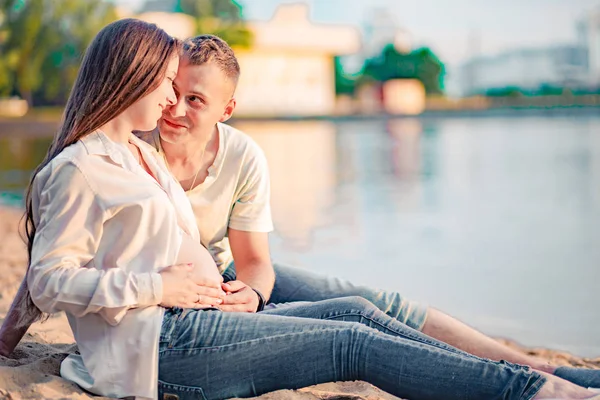 Beautiful pregnant woman and her husband are sitting on the bank of the river at sunset — Stock Photo, Image