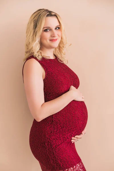Беременная женщина в бордовом платье обнимает живот о персиковую стену — стоковое фото