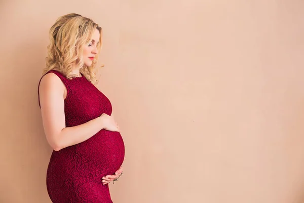 Έγκυος γυναίκα φορώντας ένα μπορντώ φόρεμα αγκαλιάζει την κοιλιά της σε ένα ροδάκινο-χρωματισμένο τοίχο — Φωτογραφία Αρχείου