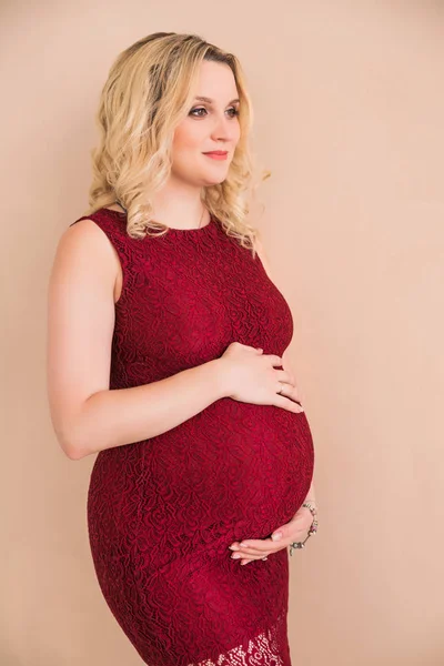 Donna incinta con un vestito bordeaux che abbraccia la pancia contro una parete color pesca — Foto Stock