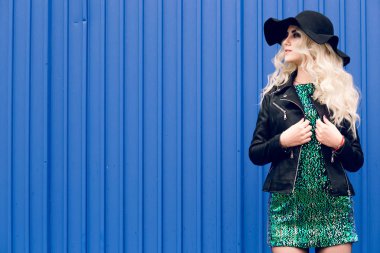 Siyah şapkalı ve deri ceketli kız duruyor ve mavi duvarın arka planına doğru poz sürüyor.