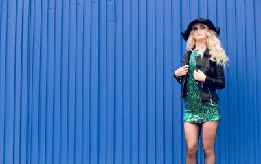 Siyah şapkalı ve deri ceketli kız duruyor ve mavi duvarın arka planına doğru poz sürüyor.