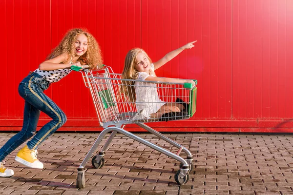 2 filles s'amusent et s'amusent avec un chariot d'épicerie sur fond de mur rouge — Photo