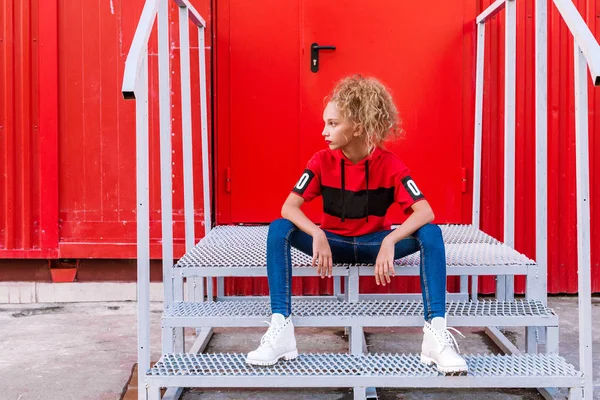Модная девушка-подросток позирует на фоне красной стены, сидит на лестнице — стоковое фото