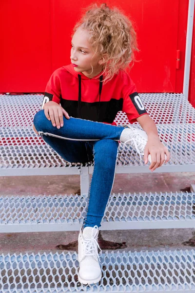 Adolescent à la mode fille posant sur un fond de mur rouge, assis sur les escaliers — Photo