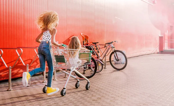 2 meninas se divertindo e se divertindo com um carrinho de supermercado contra o fundo de uma parede vermelha — Fotografia de Stock