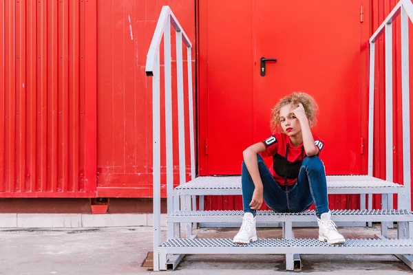 Fashionable teenager girl posing on a red wall background, sitting on the stairs — Stock Photo, Image