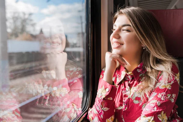 Beautiful girl sitting alone in the train and looking out the window merrily — Stock Photo, Image