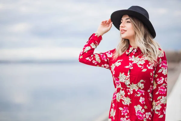 Beautiful girl in a red dress and black hat stands on the waterfront and looks into the distance — Stock Photo, Image
