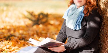 Genç kadın bir ağacın altında oturur ve sonbahar parkında bir kitap okur