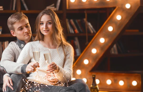 Pareja de enamorados bebe champán en una acogedora habitación con una gran lámpara en forma de carta y luces de Navidad — Foto de Stock