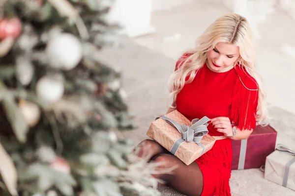Красивая девушка в красном платье сидит с рождественским подарком рядом с елкой — стоковое фото