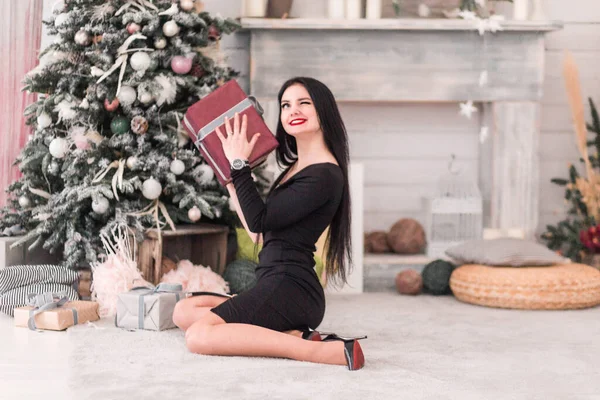 Красивая девушка в черном платье сидит с рождественским подарком рядом с елкой — стоковое фото