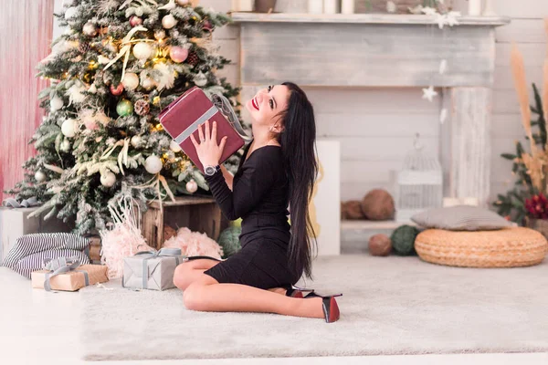 Красивая девушка в черном платье сидит с рождественским подарком рядом с елкой — стоковое фото