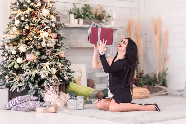 Schöne Mädchen in einem schwarzen Kleid sitzt mit einem Weihnachtsgeschenk neben dem Weihnachtsbaum — Stockfoto