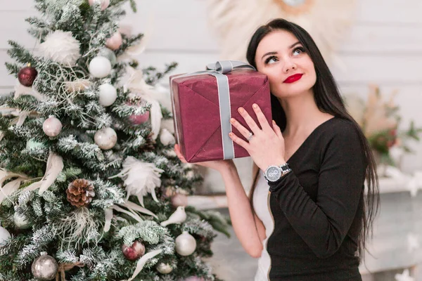 Όμορφο κορίτσι σε ένα μαύρο φόρεμα και ένα ονειρεμένο βλέμμα με ένα δώρο Πρωτοχρονιάς κοντά στο χριστουγεννιάτικο δέντρο — Φωτογραφία Αρχείου