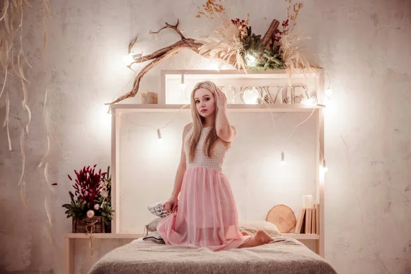 Portrait of young beautiful blond woman relax in bedroom. Smile happy caucasian girl with long curly hair wear pink dress
