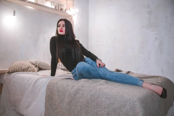 Портрет молодой красивой брюнетки женщины расслабиться в спальне. Улыбайтесь счастливая белая девушка с длинными вьющимися волосами носить теплый свитер и джинсы — стоковое фото