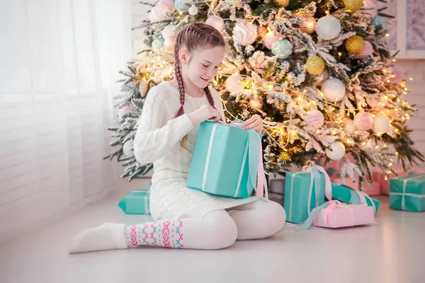 Hermosa chica se sienta en el suelo cerca del árbol de Año Nuevo y abre un regalo de Año Nuevo —  Fotos de Stock