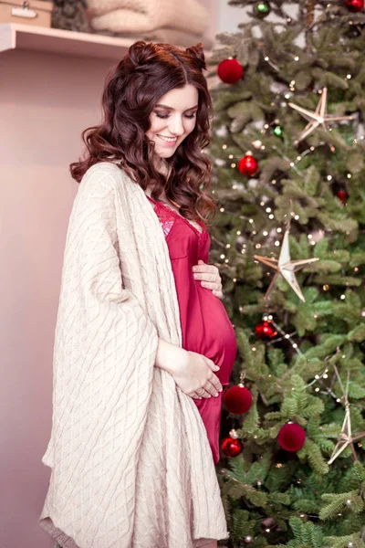 Όμορφη έγκυος γυναίκα σε ένα μπορντό πουκάμισο στέκεται κοντά στο δέντρο της Πρωτοχρονιάς — Φωτογραφία Αρχείου