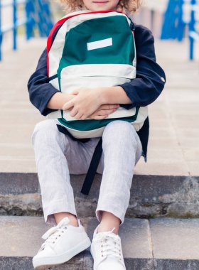 Açık renk pantolonlu ve koyu renk gömlekli bir öğrenci elinde sırt çantasıyla merdivenlerde oturuyor. Vertycal fotoğraf