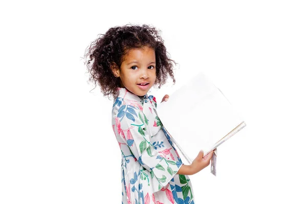 Pequeña linda chica afroamericana de moda en vestido blanco con estampado floral sosteniendo cuadernos en las manos y mirando a la cámara — Foto de Stock