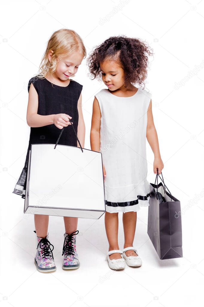 Little fashionable girls of two different nationalities are posing against a white background with bags from the store. One girl shows the other what is in her bag