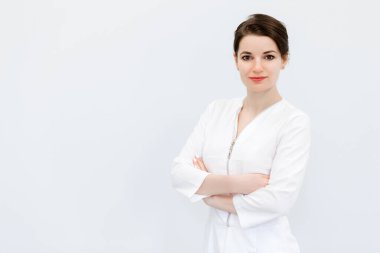 Topuz saç stili, tıbbi takım elbiseli ve beyaz bir duvara yaslanmış kollarını çaprazlamış güzel ve tatlı bir genç kadın. Yatay fotoğraf
