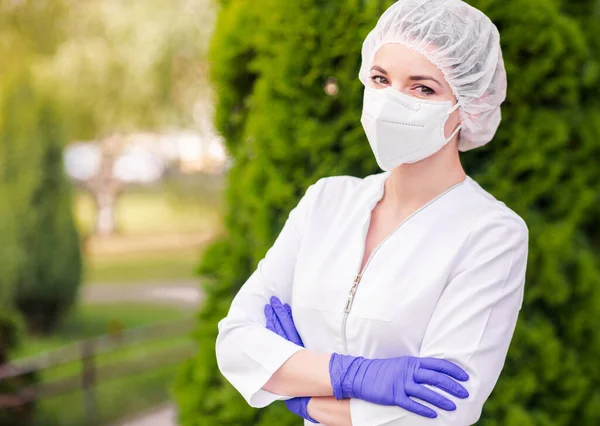 A beautiful girl in a medical suit, a protective mask on her face, gloves and a medical cap poses against a background of greenery. The girl stands with crossed arms and looks into the camera. Horizontal photo