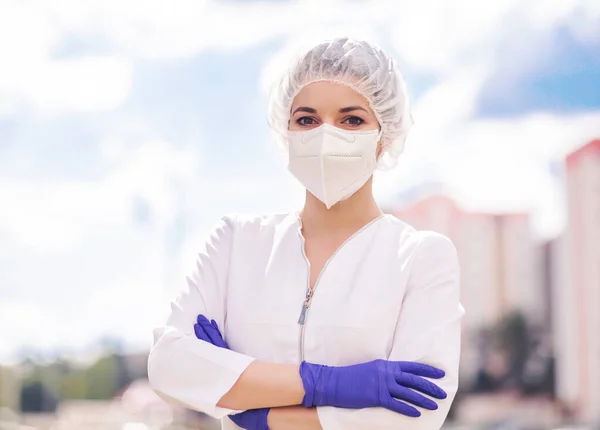 A beautiful girl in a medical suit, a protective mask on her face, gloves and a medical cap poses against the background of the blue sky, clouds and city buildings. The girl stands with crossed arms and look into the camera. The medic is protecting t