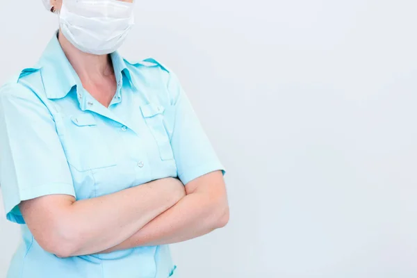 A woman in a medical suit, with a protective mask on her face and with crossed arms, poses against a white wall. Horizontal photo