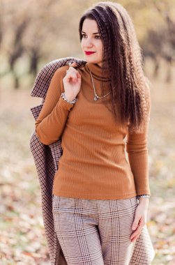 Güz parkında poz veren pantolonlu, balıkçı yakalı ve montlu güzel esmer kadın. Başka tarafa bakan bir kadın. Dikey fotoğraf