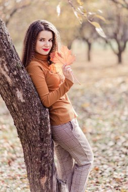 Güzel bir esmer kadın, pantolon, balıkçı yaka ve elinde bir sonbahar yaprağı ile sonbahar parkında poz veriyor. Başka tarafa bakan bir kadın. Dikey fotoğraf