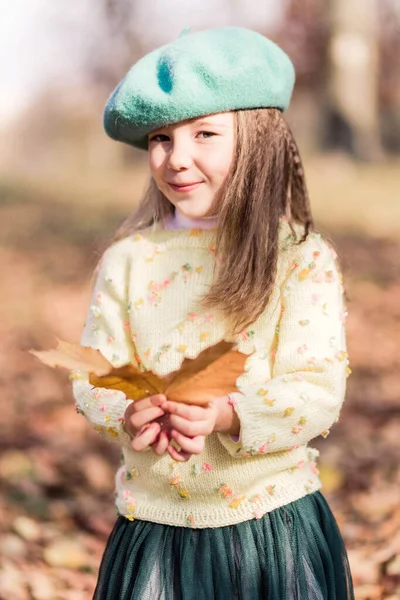 Bereli, sarı kazaklı ve sonbahar parkında yeşil tüllü etekli güzel bir kız. Elinde sonbahar sarısı bir yaprak tutan bir kız. Dikey fotoğraf