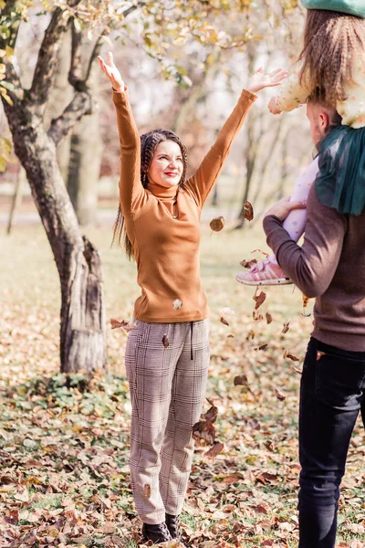 Anne, baba ve küçük kızları sonbahar parkında yürüyorlar. Kız babasının omuzlarında oturuyor. Annem sonbahar yapraklarını fırlatır. Dikey fotoğraf