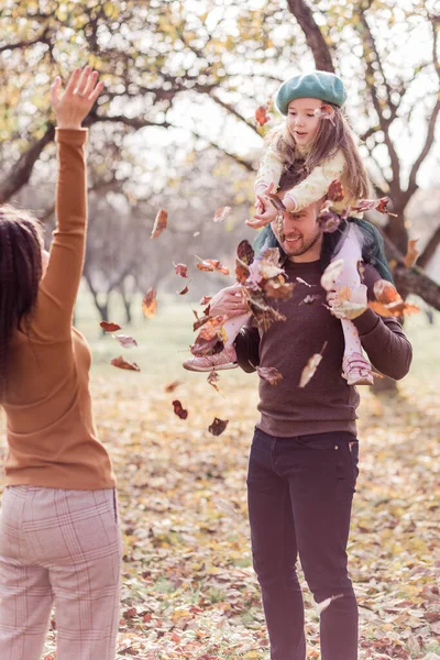 Anne, baba ve küçük kızları sonbahar parkında yürüyorlar. Kız babasının omuzlarında oturuyor. Annem sonbahar yapraklarını fırlatır. Dikey fotoğraf