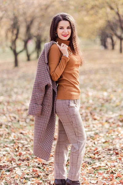 Güz parkında poz veren pantolonlu, balıkçı yakalı ve montlu güzel esmer kadın. Kadın kameraya bakıyor. Dikey fotoğraf