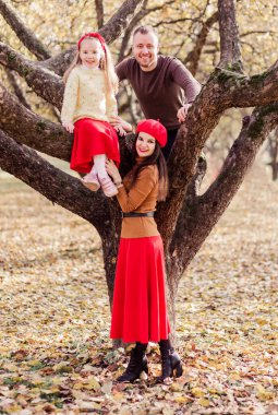 Anne, baba ve kızı parkta birlikte vakit geçiriyorlar. Baba ve kızı bir ağaca tırmandı, anne yakınlarda duruyor. Aile kameraya bakıyor. Dikey fotoğraf